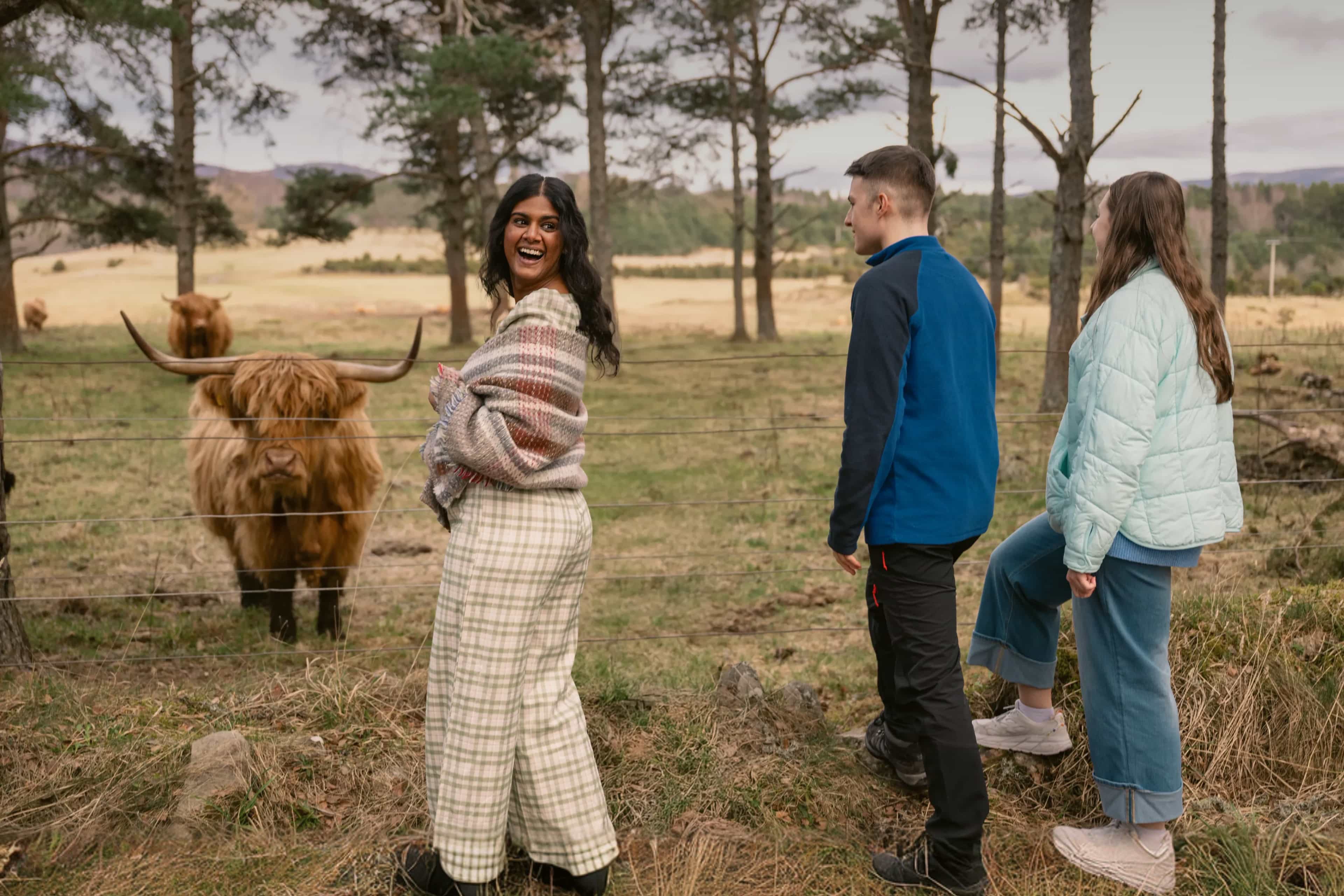 Travellers laughing while meeting a shaggy Highland cow behind a fence in the Scottish countryside.