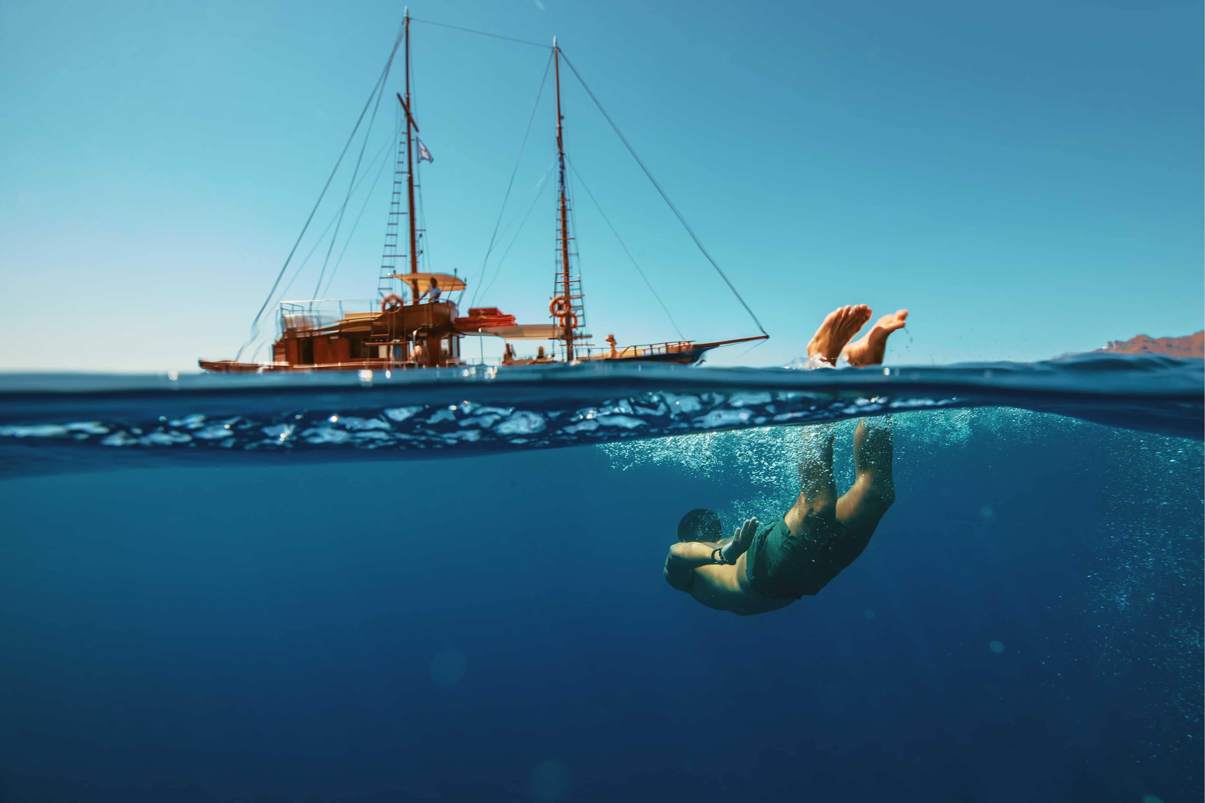 Split-view shot of a traveller diving underwater beside a wooden sailing boat in clear blue sea in Greece.