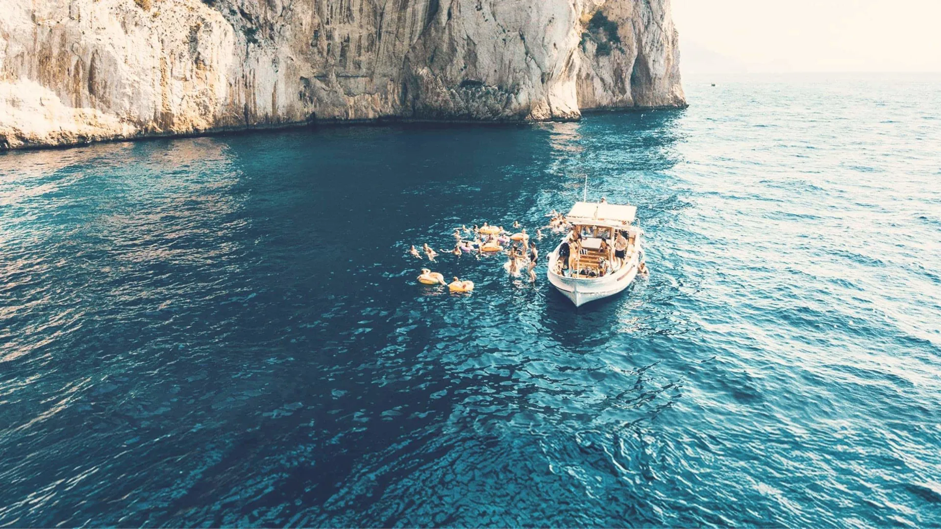 Contiki travellers swimming from a boat beneath the towering cliffs of Capri, Italy, on the Ultimate European Plus Greek Islands Tour