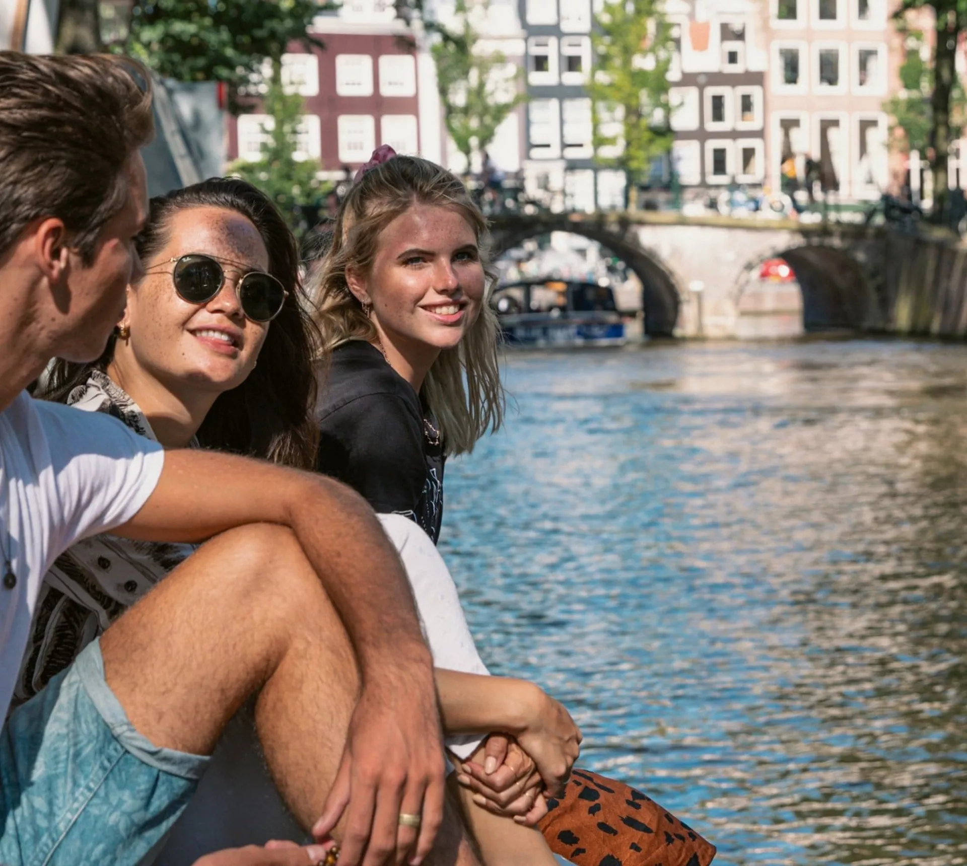 Contiki travellers relax beside an Amsterdam canal, with historic bridges and canal houses in the background on the European Escapade Tour