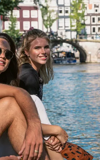 Contiki travellers relax beside an Amsterdam canal, with historic bridges and canal houses in the background on the European Escapade Tour