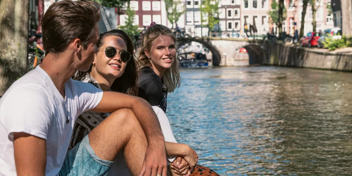 Contiki travellers relax beside an Amsterdam canal, with historic bridges and canal houses in the background on the European Escapade Tour