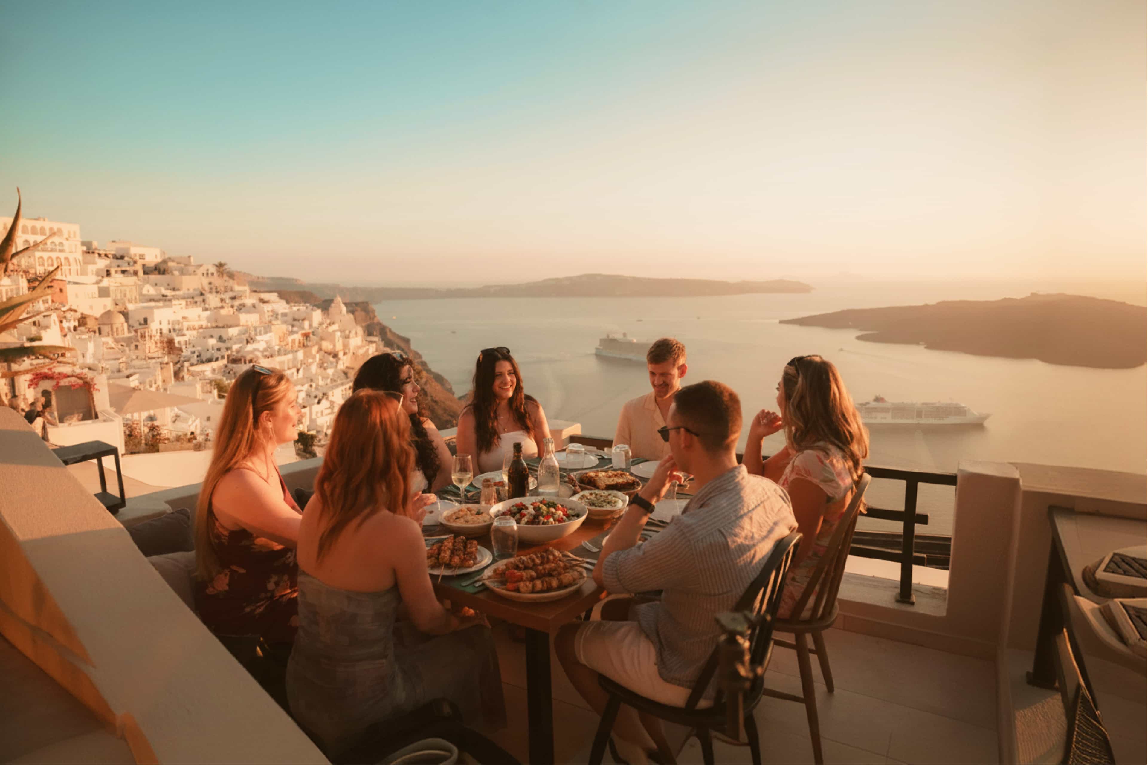 Travellers sharing a Greek meal on a clifftop terrace overlooking the caldera at sunset in Santorini, Greece.