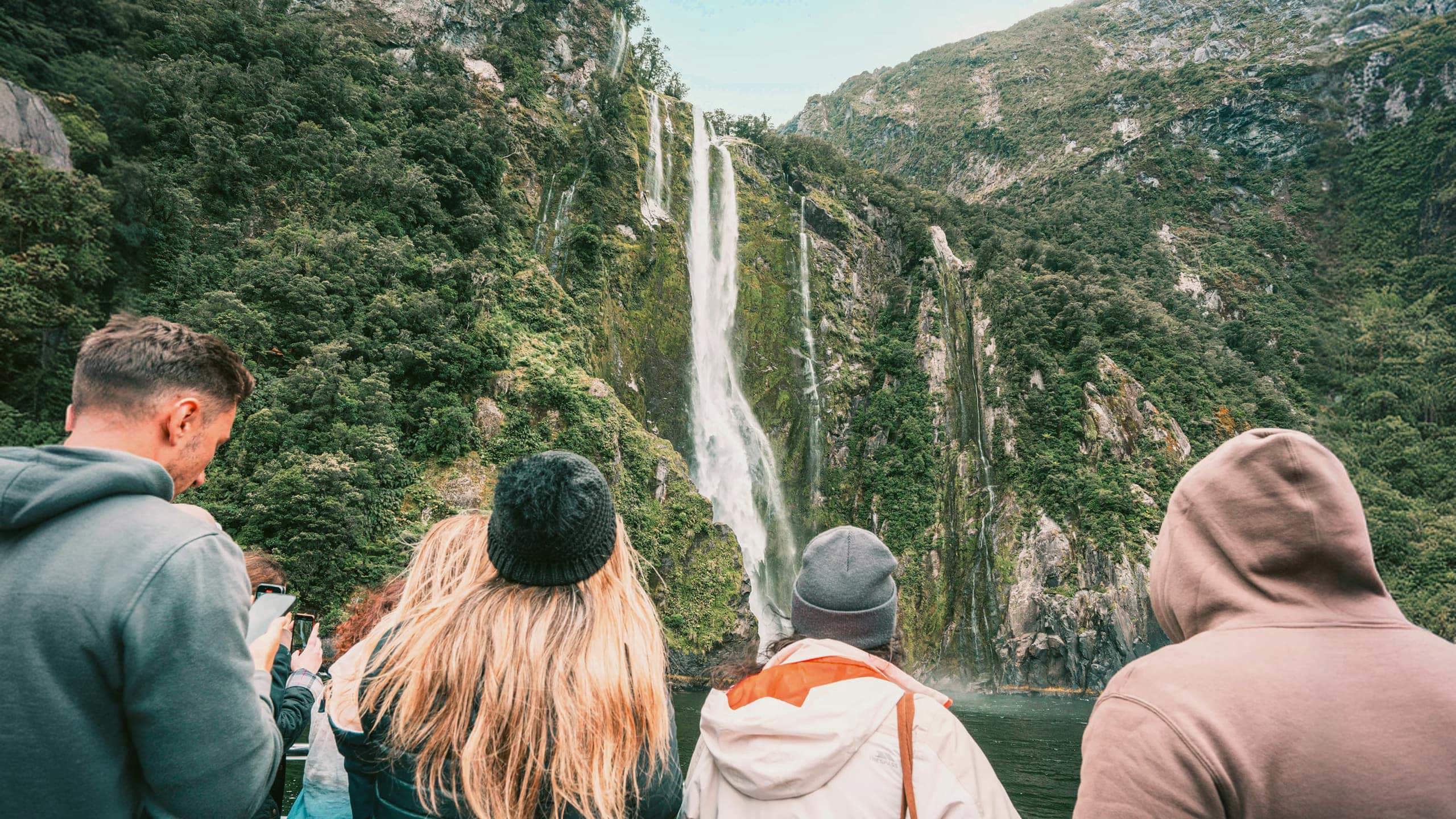 Travellers on a Milford Sound cruise watching a waterfall cascade down lush rainforest fiord walls in Fiordland National Park New Zealand - Contiki South Island Tour