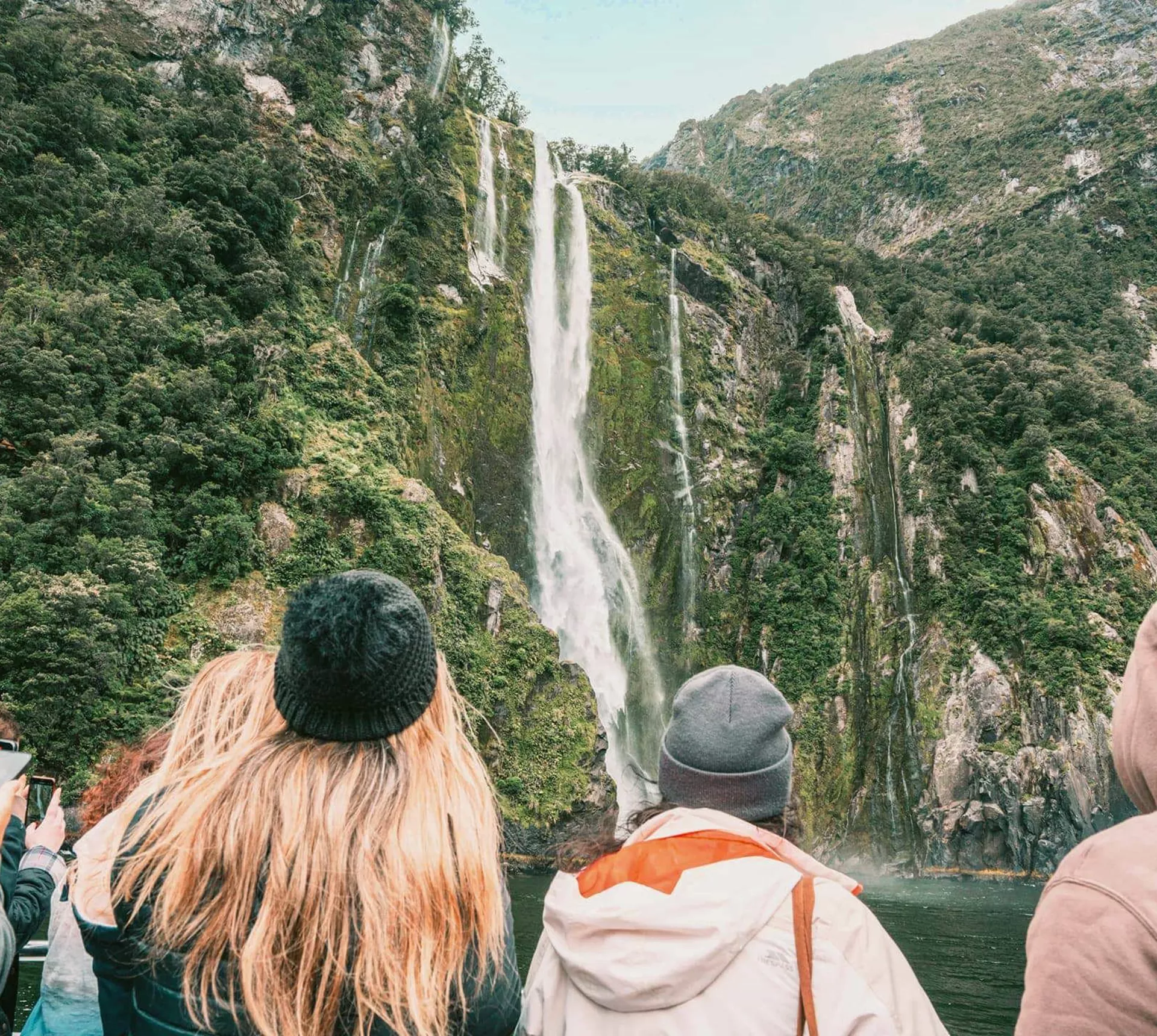 Travellers on a Milford Sound cruise watching a waterfall cascade down lush rainforest fiord walls in Fiordland National Park New Zealand - Contiki South Island Tour