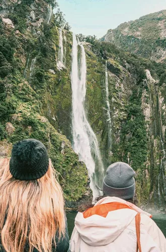 Travellers on a Milford Sound cruise watching a waterfall cascade down lush rainforest fiord walls in Fiordland National Park New Zealand - Contiki South Island Tour