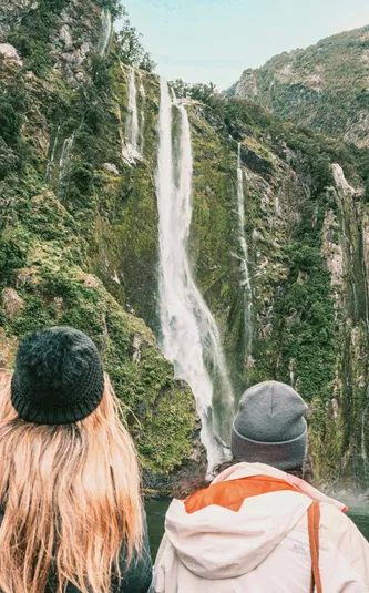 Travellers on a Milford Sound cruise watching a waterfall cascade down lush rainforest fiord walls in Fiordland National Park New Zealand - Contiki South Island Tour