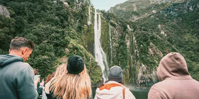 Travellers on a Milford Sound cruise watching a waterfall cascade down lush rainforest fiord walls in Fiordland National Park New Zealand - Contiki South Island Tour