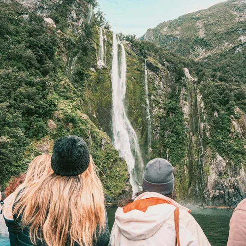 Travellers on a Milford Sound cruise watching a waterfall cascade down lush rainforest fiord walls in Fiordland National Park New Zealand - Contiki South Island Tour