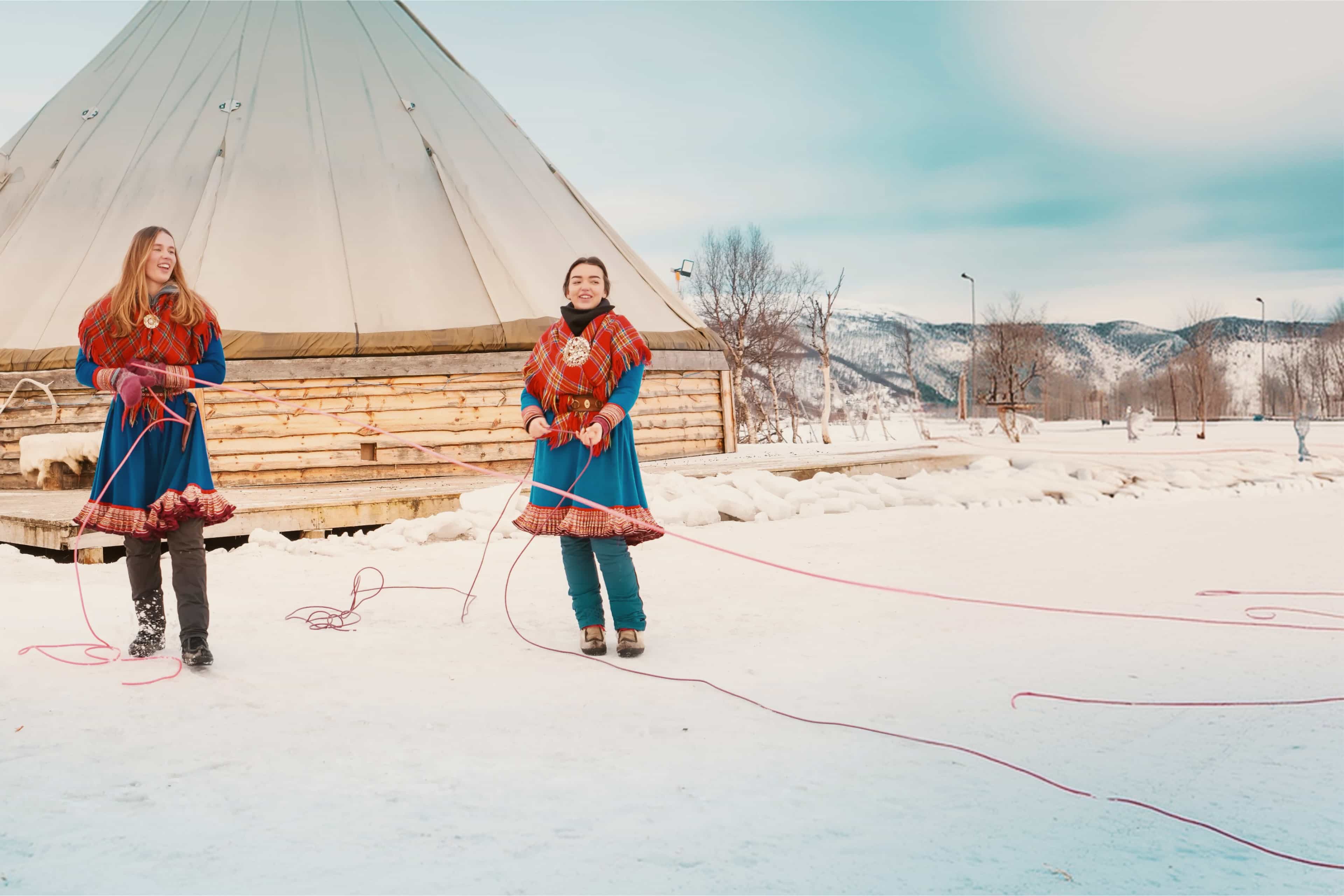 Travellers in traditional Sámi dress practising lasso throwing beside a lavvu tent in snowy Norway.