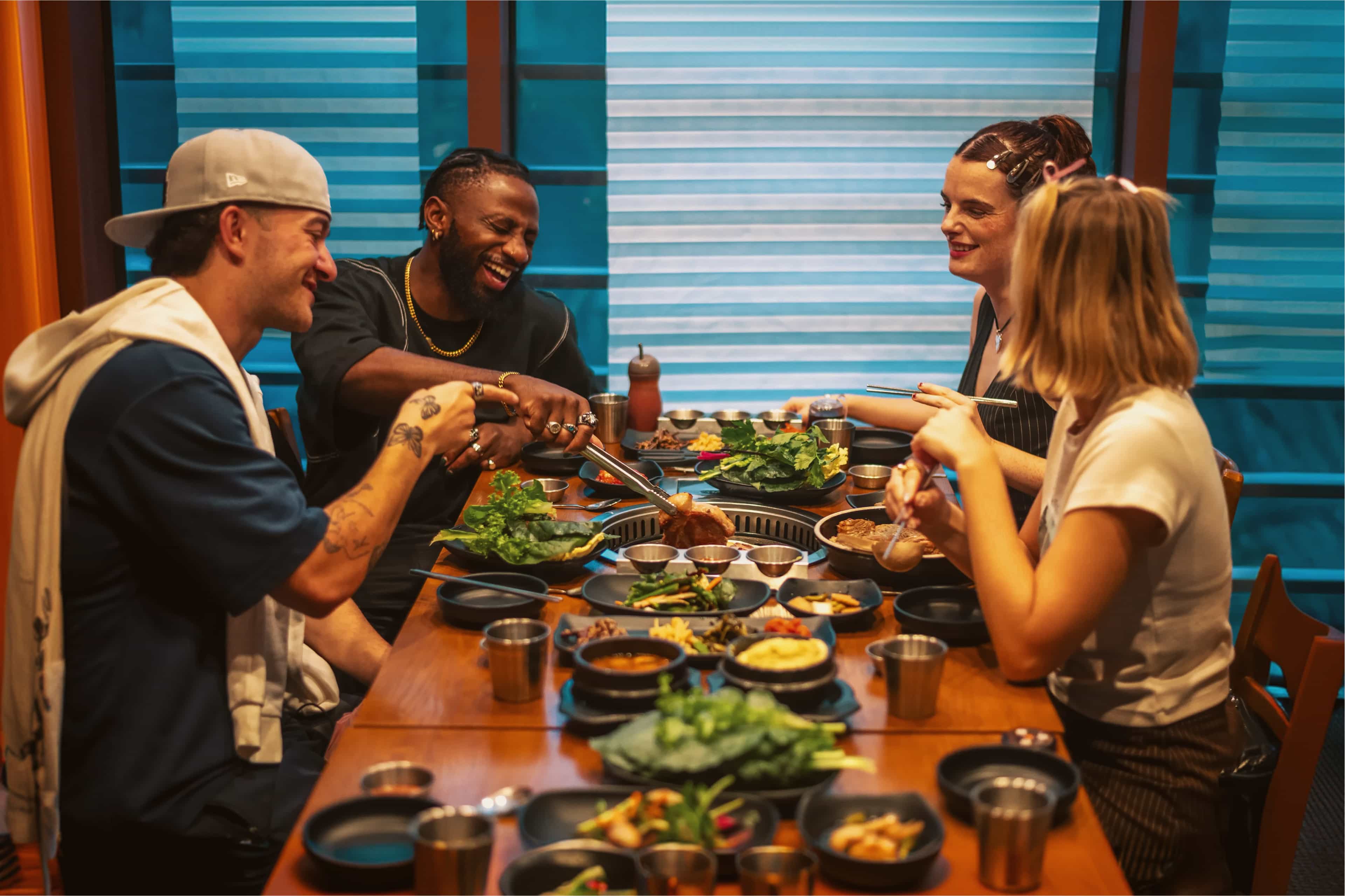 Friends laugh and share a Korean BBQ meal together, grilling meat and enjoying a spread of banchan side dishes.