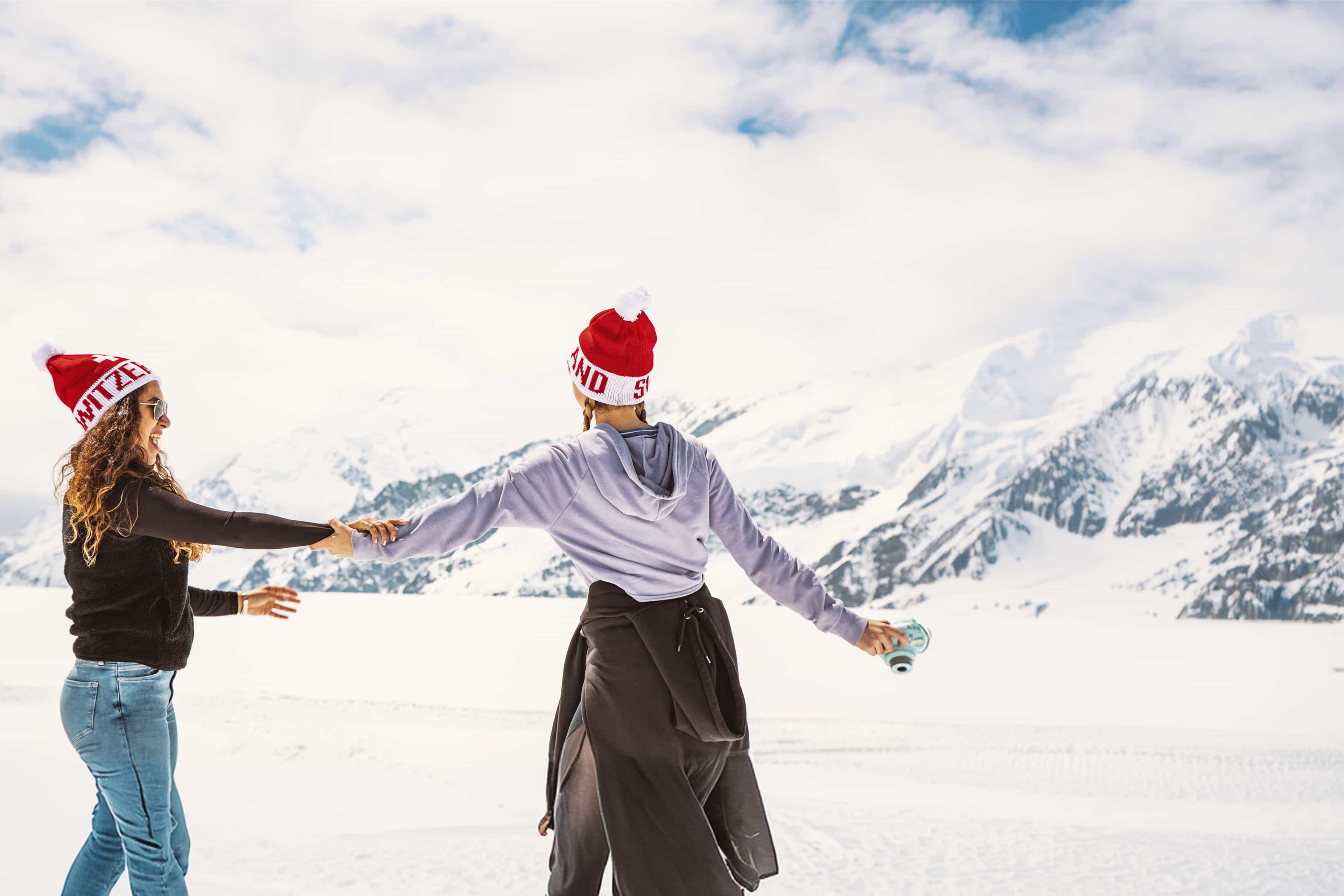 Two travellers in Switzerland beanies holding hands on a snow-covered glacier with mountains behind, Switzerland.