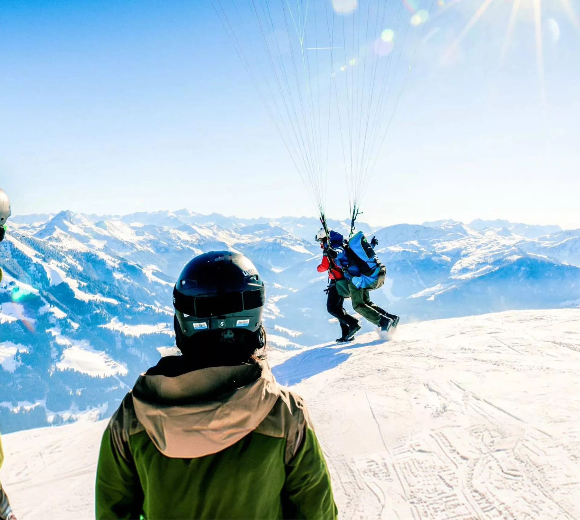 Two skiers in helmets watching a paraglider launch from a snowy Alpine summit with breathtaking panoramic views of the Austrian Alps mountain range - Contiki Ski Austria Tour