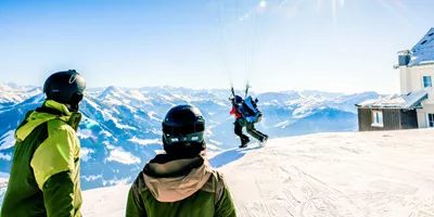 Two skiers in helmets watching a paraglider launch from a snowy Alpine summit with breathtaking panoramic views of the Austrian Alps mountain range - Contiki Ski Austria Tour