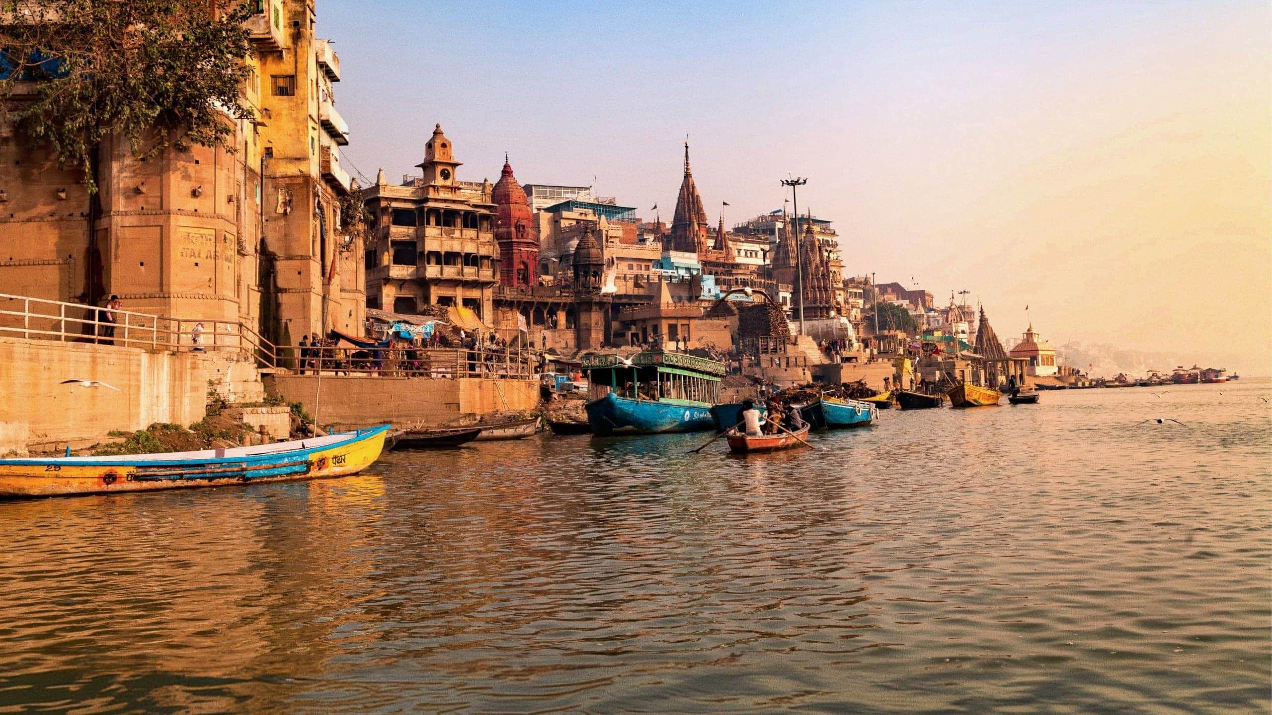 Boats drift along the Ganges River at sunrise, with the historic ghats and temples of Varanasi lining the waterfront - Delhi to Varanasi by Train Tour