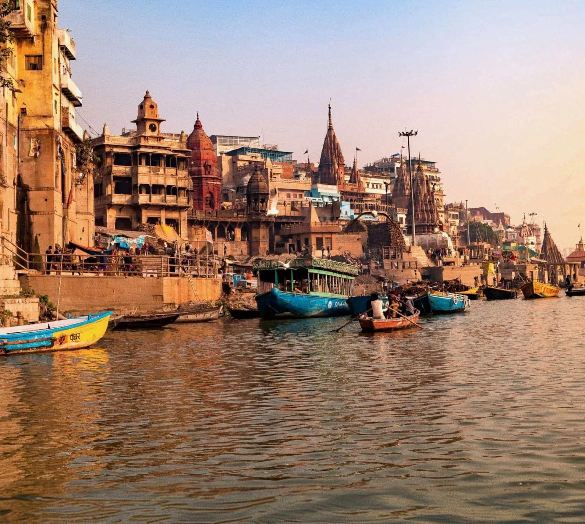 Boats drift along the Ganges River at sunrise, with the historic ghats and temples of Varanasi lining the waterfront - Delhi to Varanasi by Train Tour