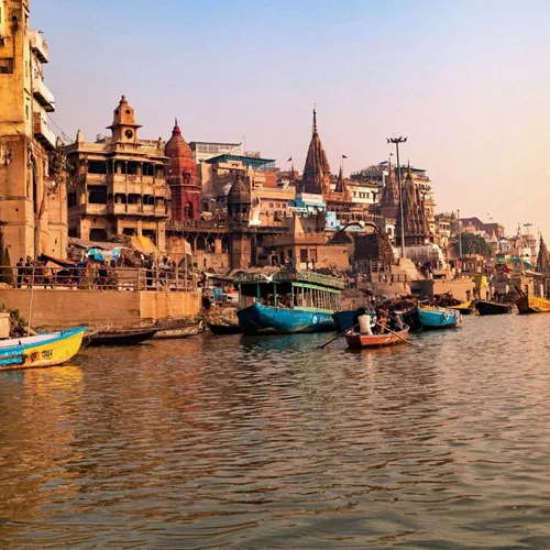 Boats drift along the Ganges River at sunrise, with the historic ghats and temples of Varanasi lining the waterfront - Delhi to Varanasi by Train Tour