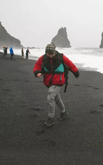 Traveller walks along Reynisfjara Black Sand Beach with the Reynisdrangar sea stacks in Iceland on the Contiki Best of Iceland Tour