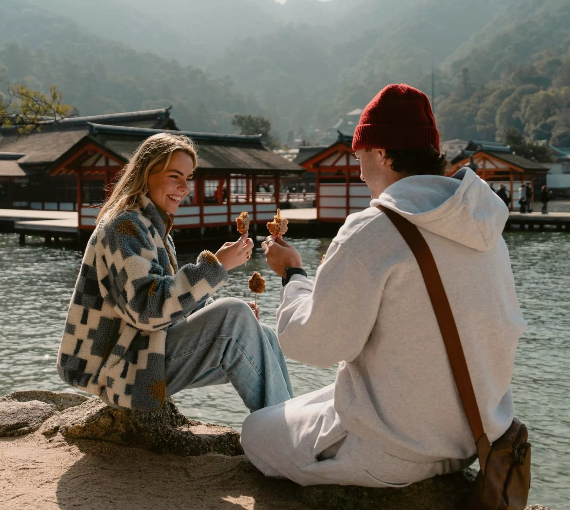 Two travellers enjoy grilled street food beside the waterfront on Miyajima Island, Japan, with the iconic Itsukushima Shrine and forested mountains in the background.