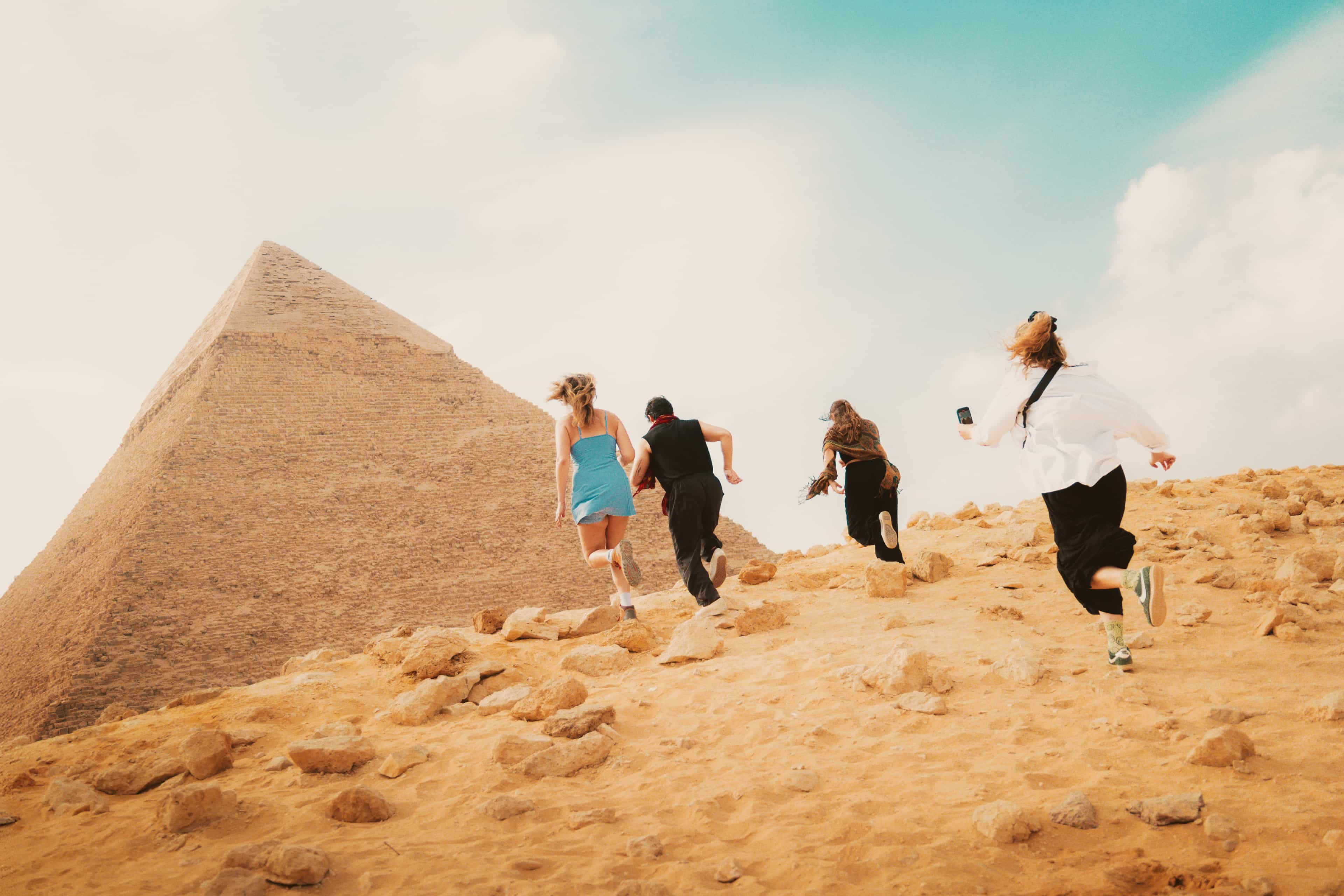 A group of young travellers run joyfully up a sandy, rock-strewn hill toward the Great Pyramid of Giza under a warm, hazy sky.
