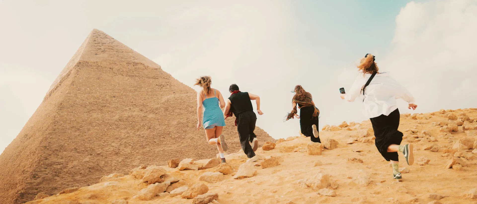 A group of young travellers run joyfully up a sandy, rock-strewn hill toward the Great Pyramid of Giza under a warm, hazy sky.