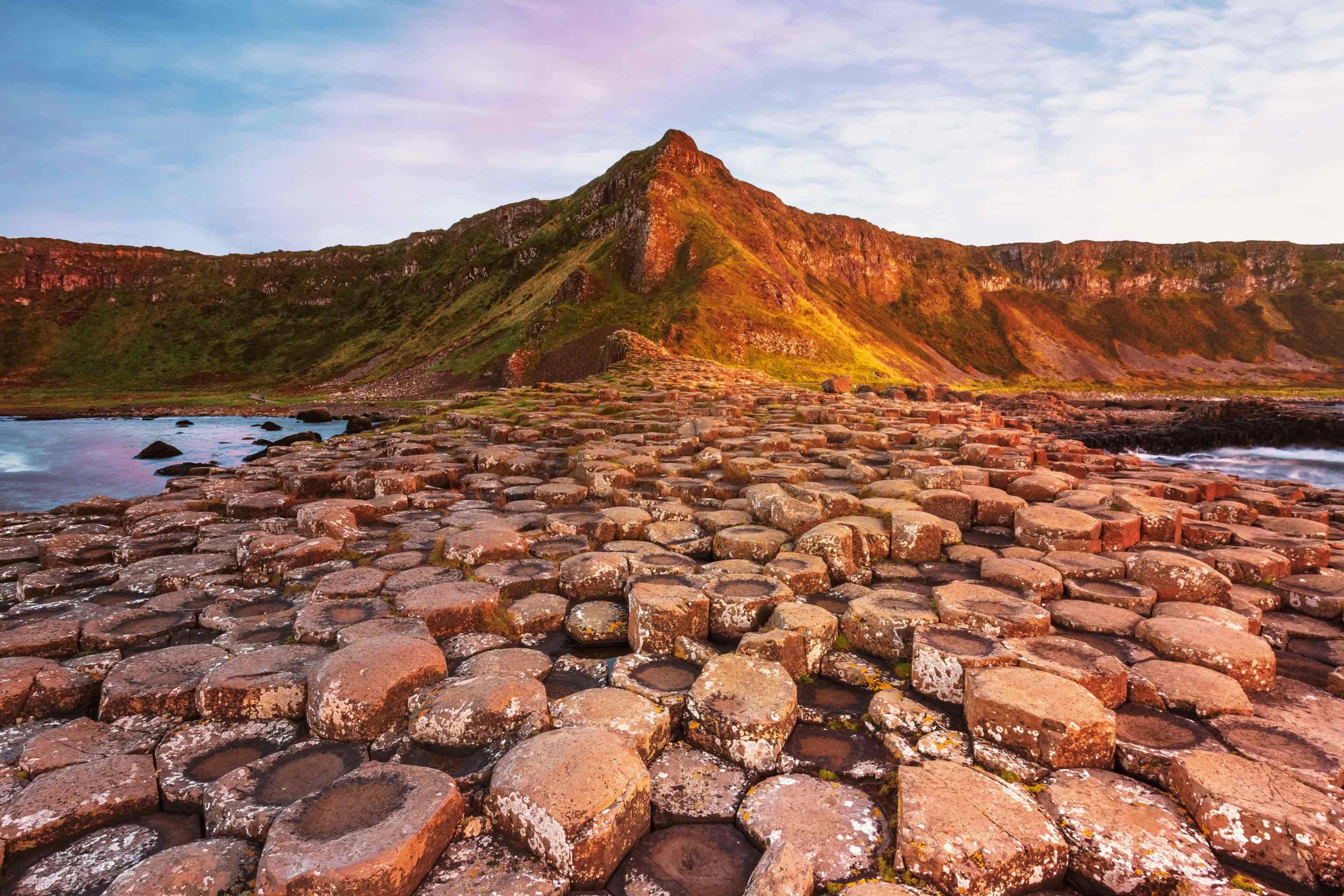 Hexagonal basalt columns of the Giant's Causeway leading toward green cliffs at sunset, Northern Ireland.