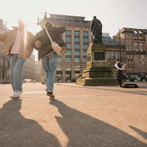 Travellers explore George Square in Glasgow while a bagpiper performs on the Contiki Best of England & Scotland Tour
