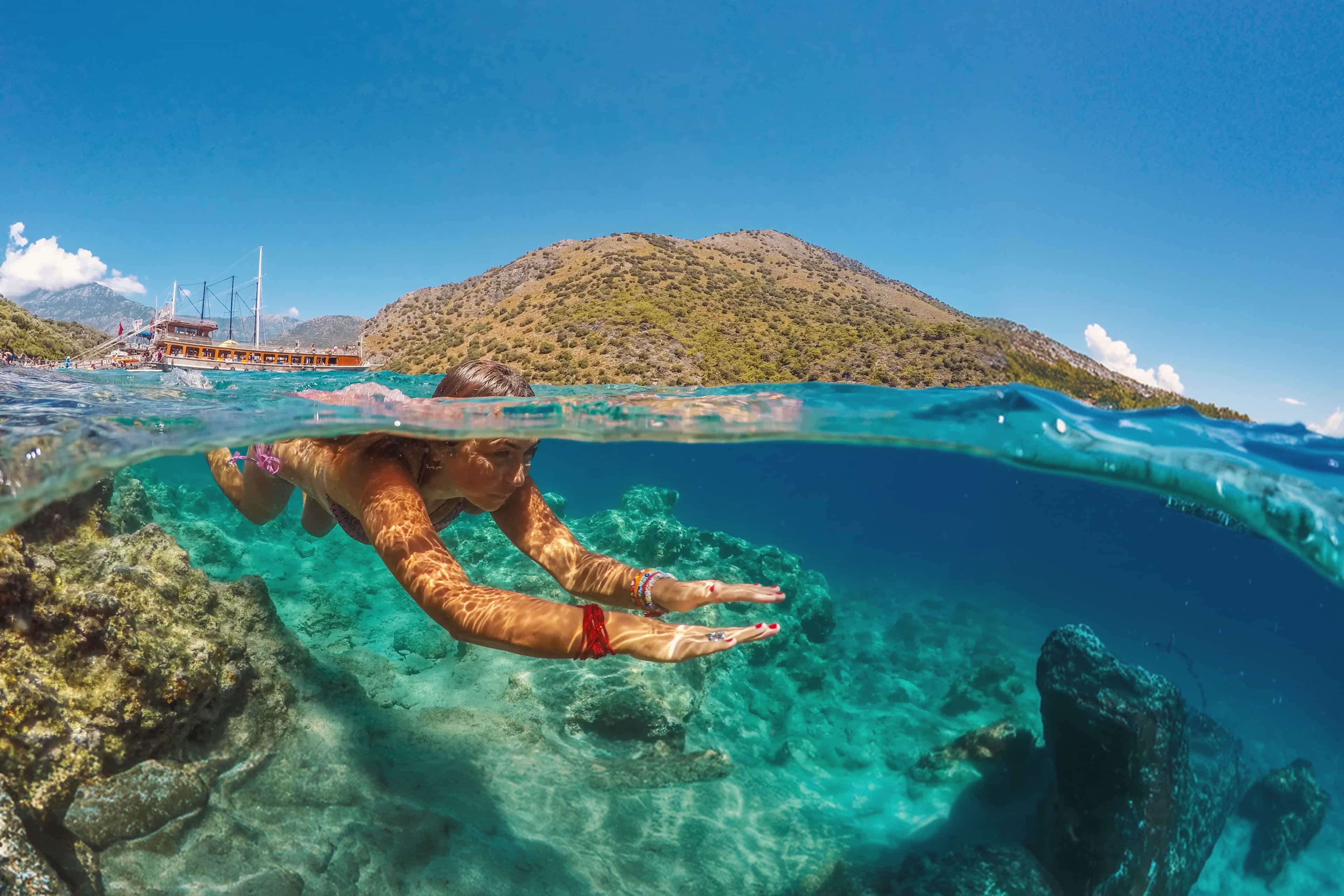 Split-view shot of a traveller swimming over rocky reef in clear turquoise water, with a gulet boat and hills behind, Turkey.