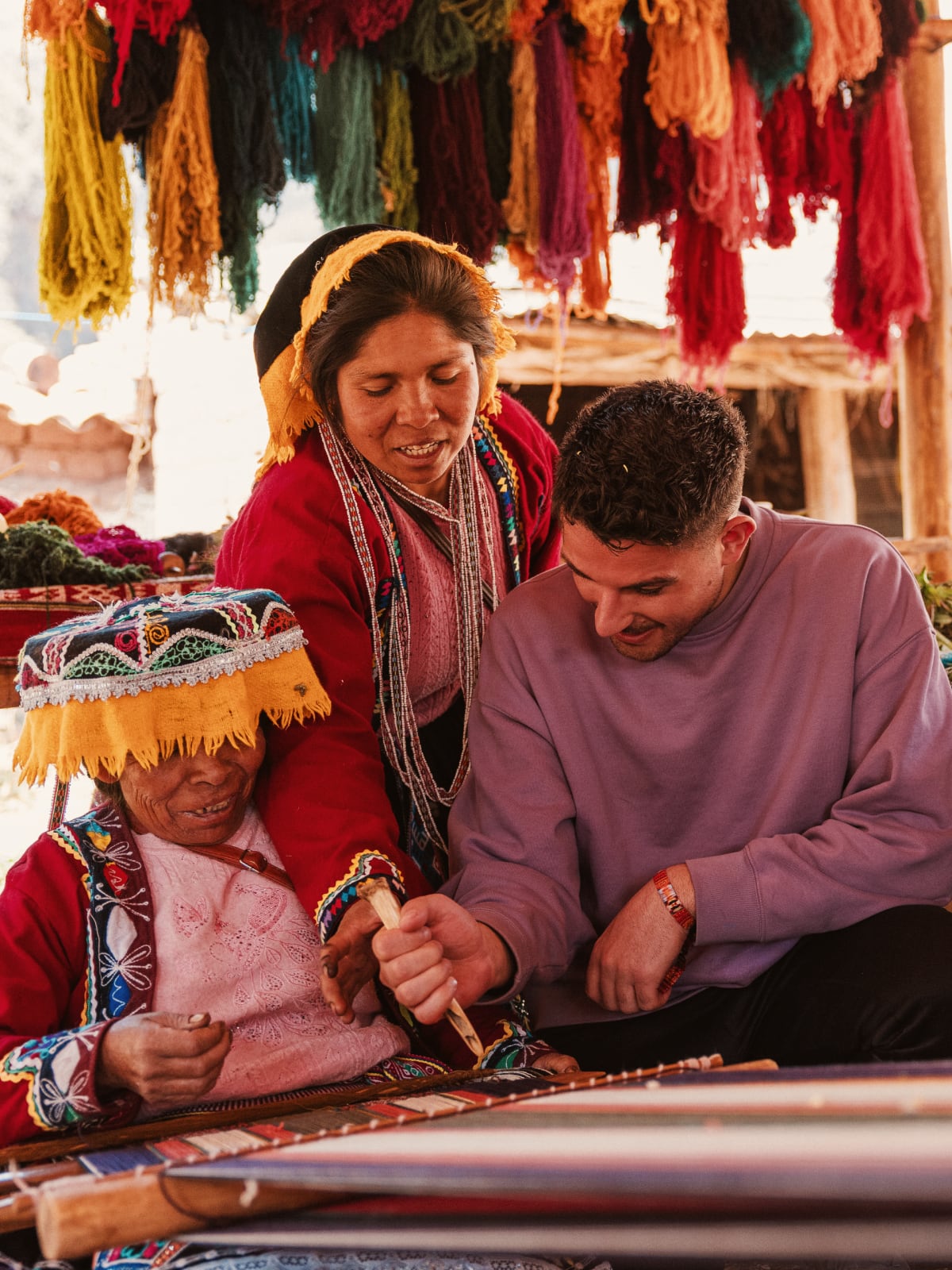 Local weavers in traditional dress teaching a traveller hand-weaving techniques beneath dyed wool skeins in Peru.