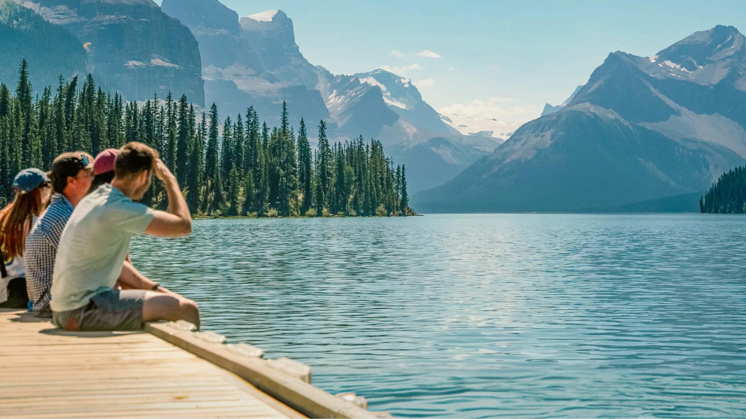 Three travellers sitting on a wooden dock at Maligne Lake in Jasper National Park overlooking Spirit Island and the dramatic Canadian Rocky Mountain peaks in Alberta - Contiki Canada Vancouver and the Rockies Tour