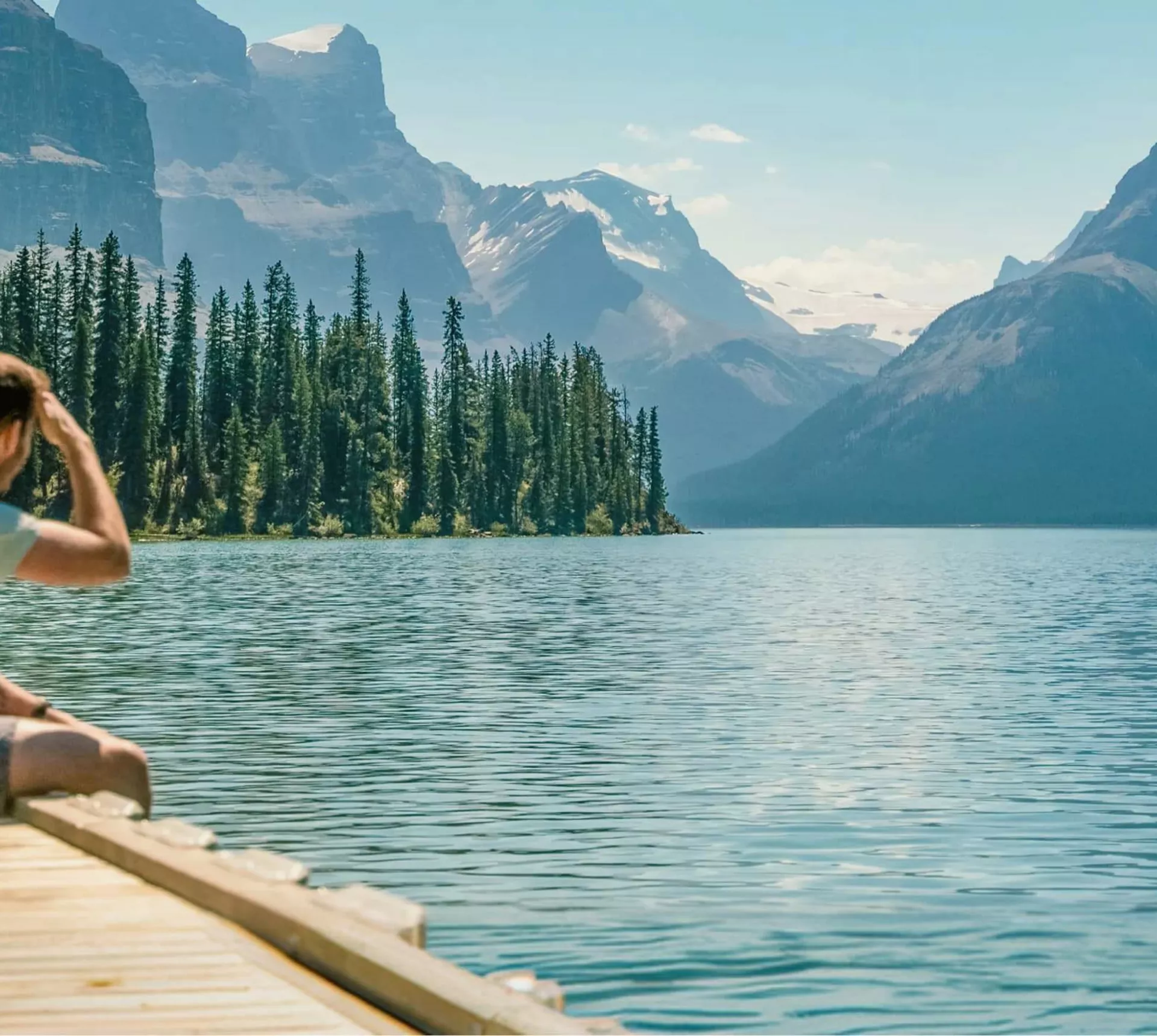 Three travellers sitting on a wooden dock at Maligne Lake in Jasper National Park overlooking Spirit Island and the dramatic Canadian Rocky Mountain peaks in Alberta - Contiki Canada Vancouver and the Rockies Tour