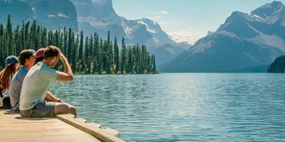 Three travellers sitting on a wooden dock at Maligne Lake in Jasper National Park overlooking Spirit Island and the dramatic Canadian Rocky Mountain peaks in Alberta - Contiki Canada Vancouver and the Rockies Tour