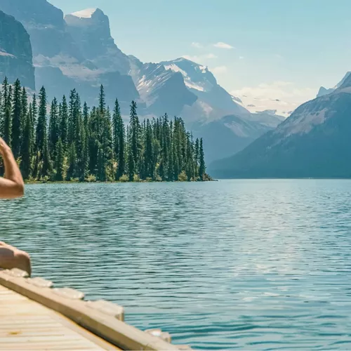 Three travellers sitting on a wooden dock at Maligne Lake in Jasper National Park overlooking Spirit Island and the dramatic Canadian Rocky Mountain peaks in Alberta - Contiki Canada Vancouver and the Rockies Tour
