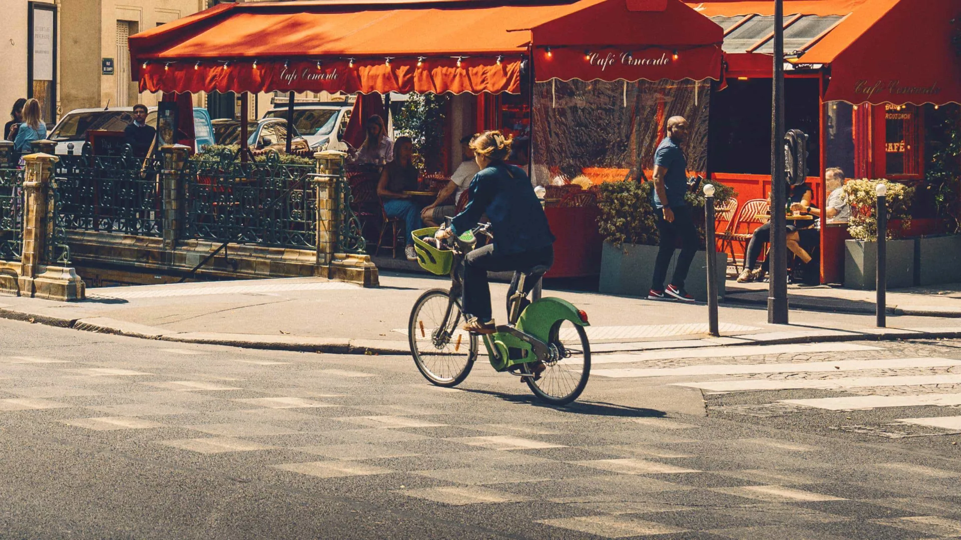Woman cycling on a bike past a classic red-awninged French café terrace on a sunny Paris street - Contiki Europe Tour