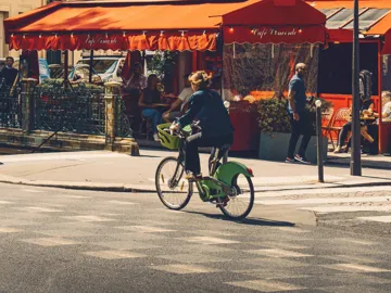 Woman cycling on a bike past a classic red-awninged French café terrace on a sunny Paris street - Contiki Europe Tour