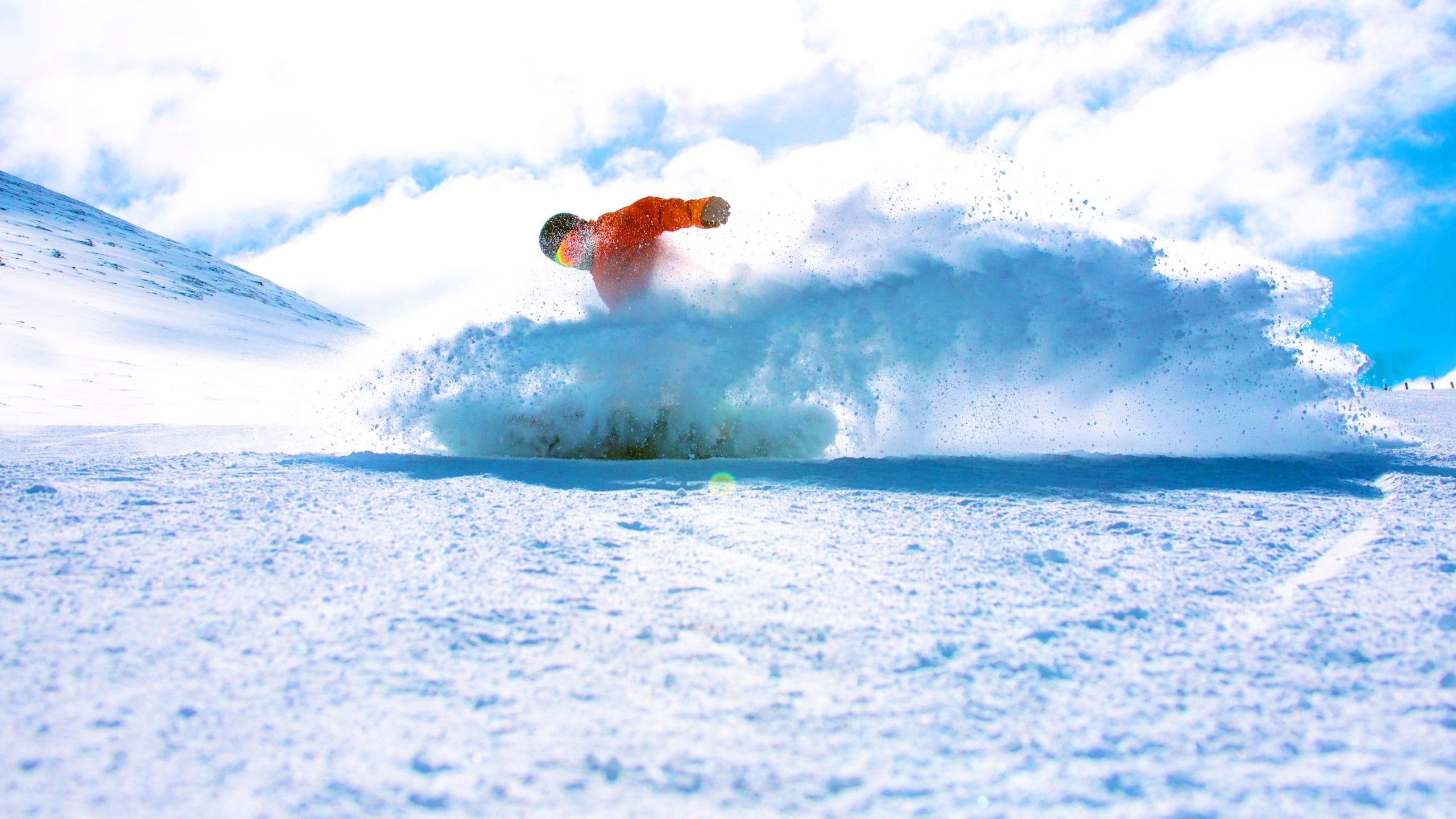 A snowboarder in a bright orange jacket rides through deep powder on a Canadian mountain, kicking up a spray of snow beneath a blue sky - Contiki Ski Canada Tour