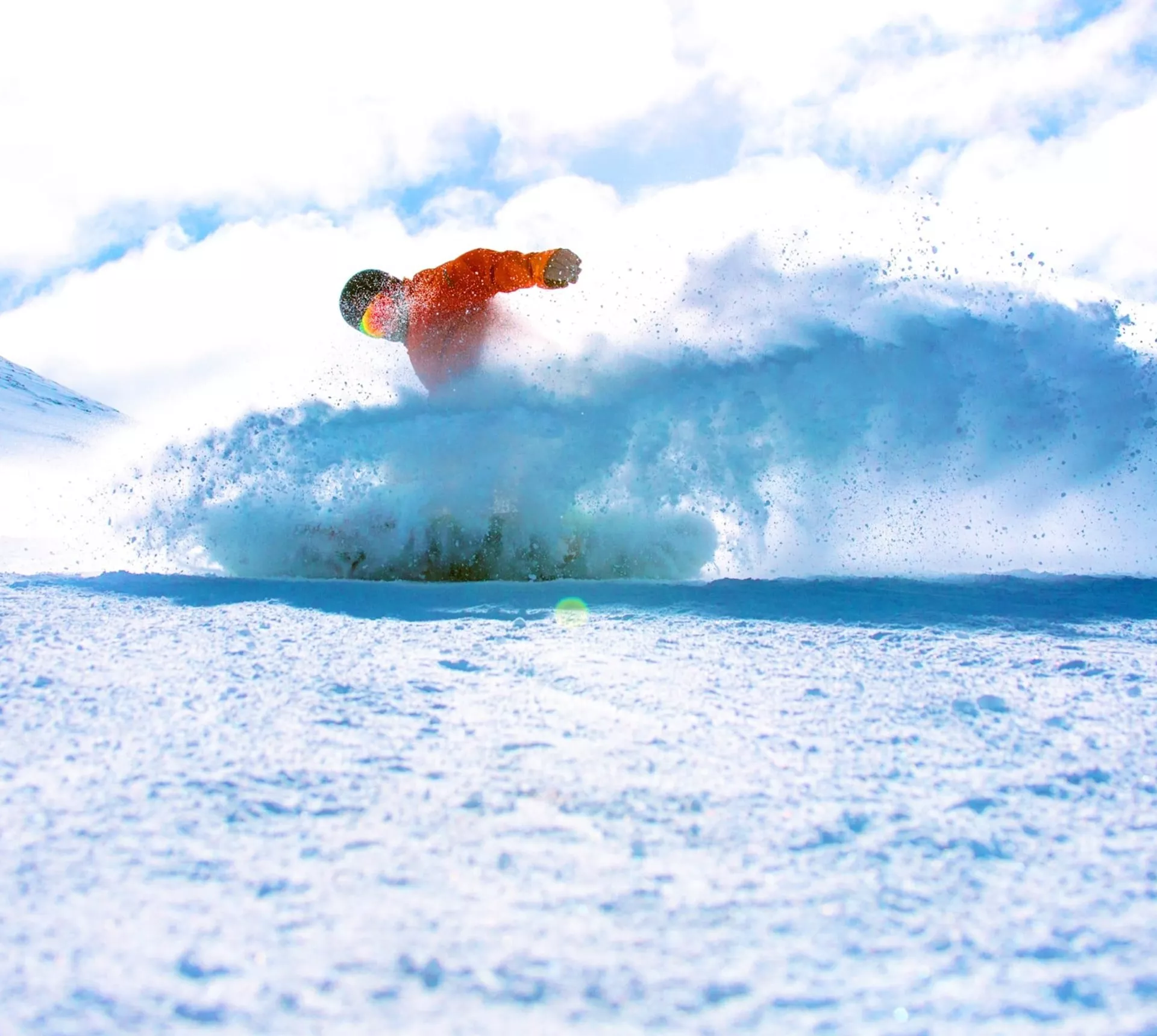 A snowboarder in a bright orange jacket rides through deep powder on a Canadian mountain, kicking up a spray of snow beneath a blue sky - Contiki Ski Canada Tour