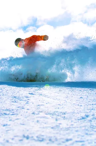 A snowboarder in a bright orange jacket rides through deep powder on a Canadian mountain, kicking up a spray of snow beneath a blue sky - Contiki Ski Canada Tour