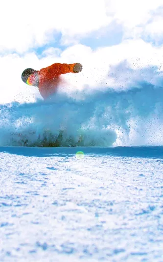 A snowboarder in a bright orange jacket rides through deep powder on a Canadian mountain, kicking up a spray of snow beneath a blue sky - Contiki Ski Canada Tour