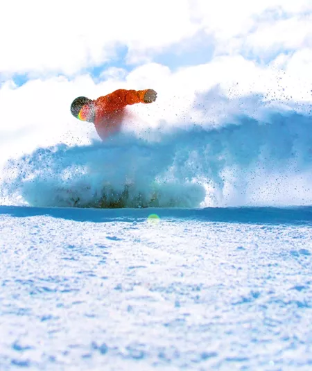 A snowboarder in a bright orange jacket rides through deep powder on a Canadian mountain, kicking up a spray of snow beneath a blue sky - Contiki Ski Canada Tour