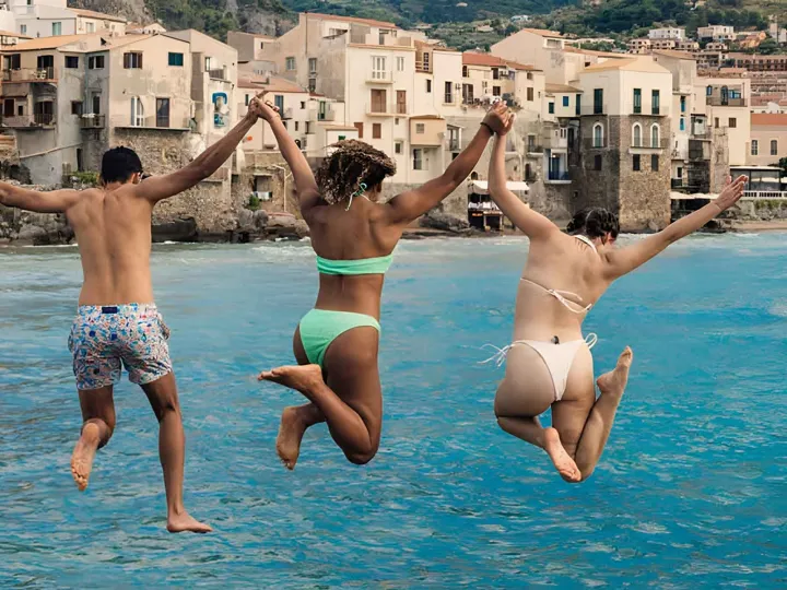 Three Young Travelers Jumping Into Sea In Sicily