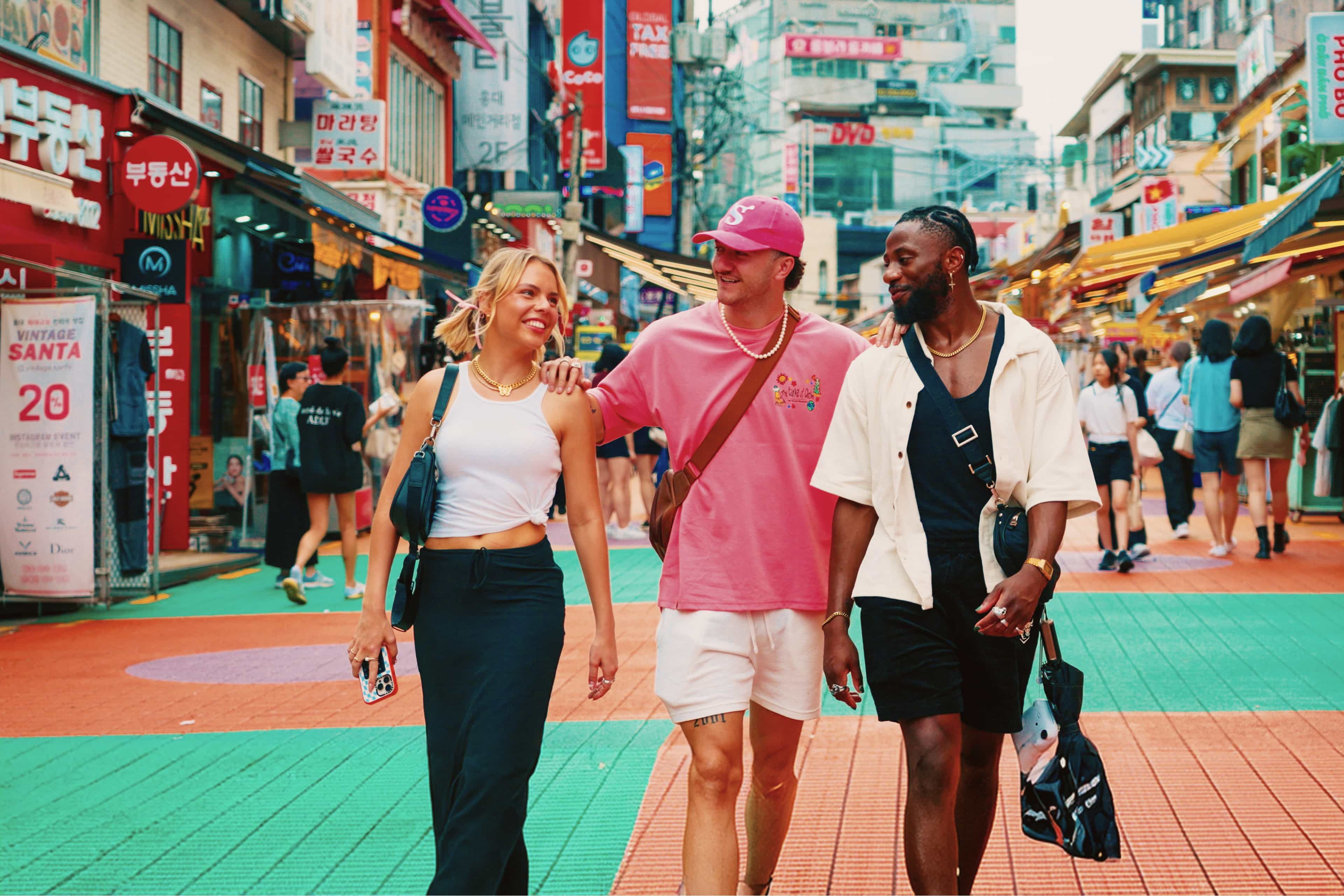 Three friends walk arm in arm through a colourful, sign-lined shopping street in Seoul, South Korea.