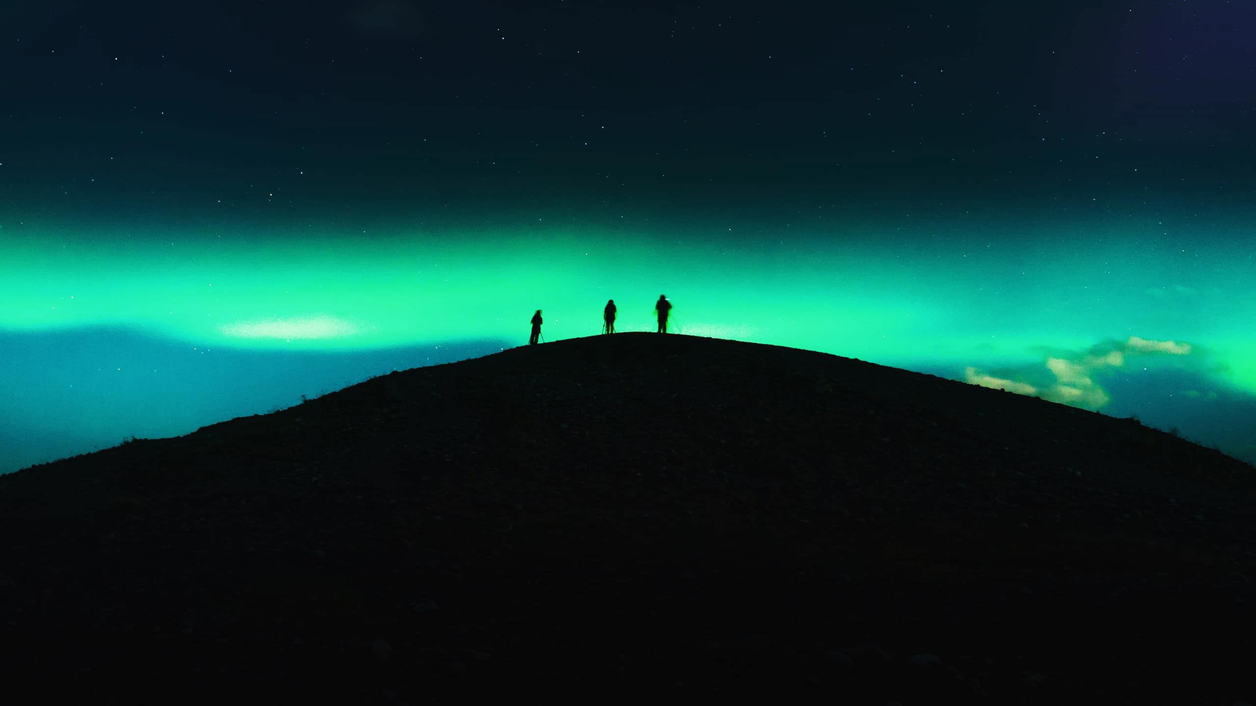 Three travellers silhouetted on a dark hilltop beneath a vivid green Aurora Borealis and starry night sky in Tromsø Norway - Contiki Norway Fjords and Northern Lights Tour