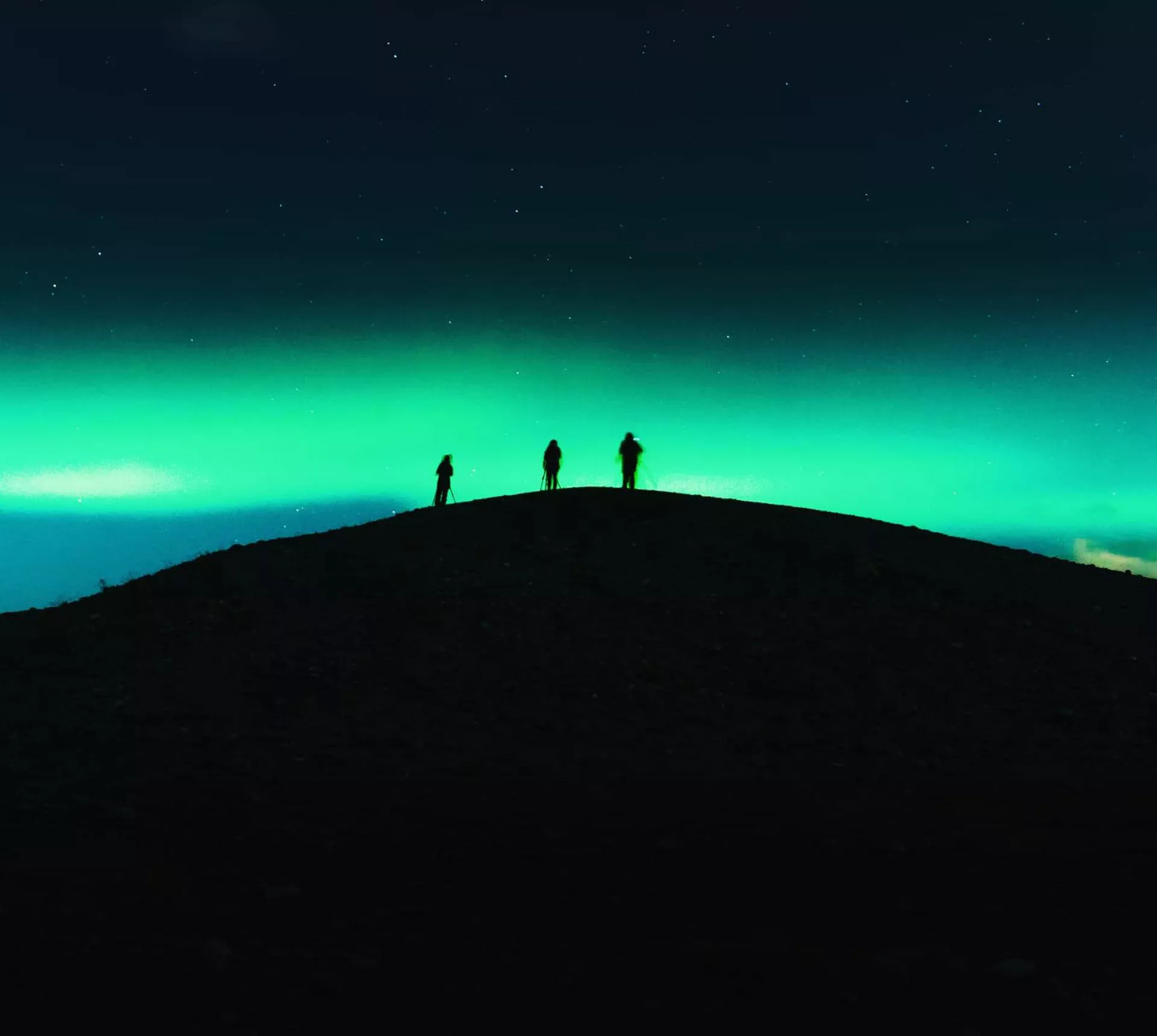 Three travellers silhouetted on a dark hilltop beneath a vivid green Aurora Borealis and starry night sky in Tromsø Norway - Contiki Norway Fjords and Northern Lights Tour
