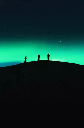 Three travellers silhouetted on a dark hilltop beneath a vivid green Aurora Borealis and starry night sky in Tromsø Norway - Contiki Norway Fjords and Northern Lights Tour