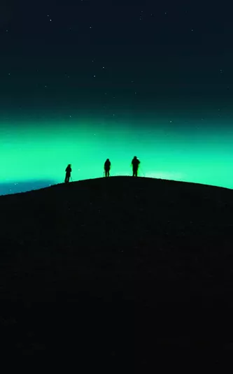 Three travellers silhouetted on a dark hilltop beneath a vivid green Aurora Borealis and starry night sky in Tromsø Norway - Contiki Norway Fjords and Northern Lights Tour