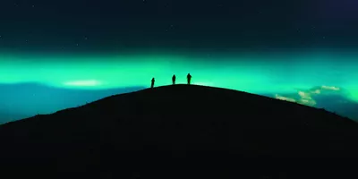 Three travellers silhouetted on a dark hilltop beneath a vivid green Aurora Borealis and starry night sky in Tromsø Norway - Contiki Norway Fjords and Northern Lights Tour