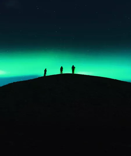 Three travellers silhouetted on a dark hilltop beneath a vivid green Aurora Borealis and starry night sky in Tromsø Norway - Contiki Norway Fjords and Northern Lights Tour