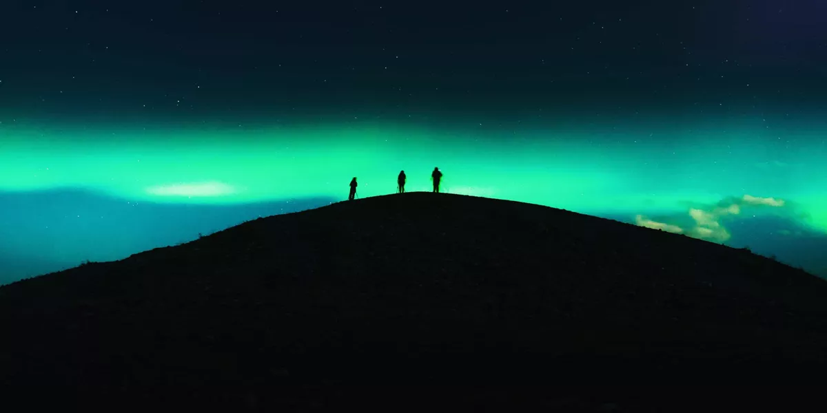 Three travellers silhouetted on a dark hilltop beneath a vivid green Aurora Borealis and starry night sky in Tromsø Norway - Contiki Norway Fjords and Northern Lights Tour
