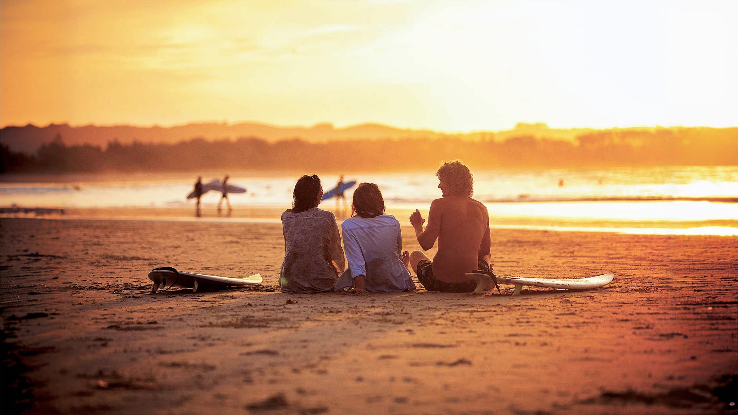 Contiki travellers relaxing on the sand with surfboards watching surfers walk along the shoreline at a stunning golden sunset in Byron Bay Australia